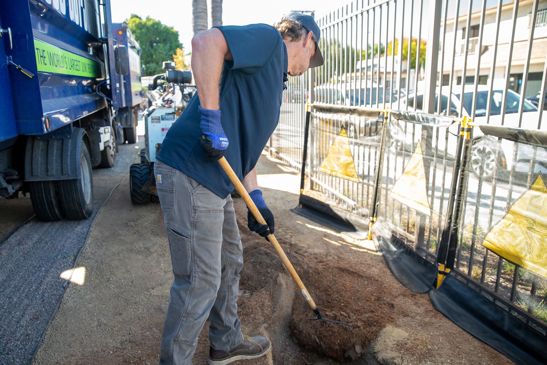 Worker digging a trench beside a truck and metal fence on a sunny street