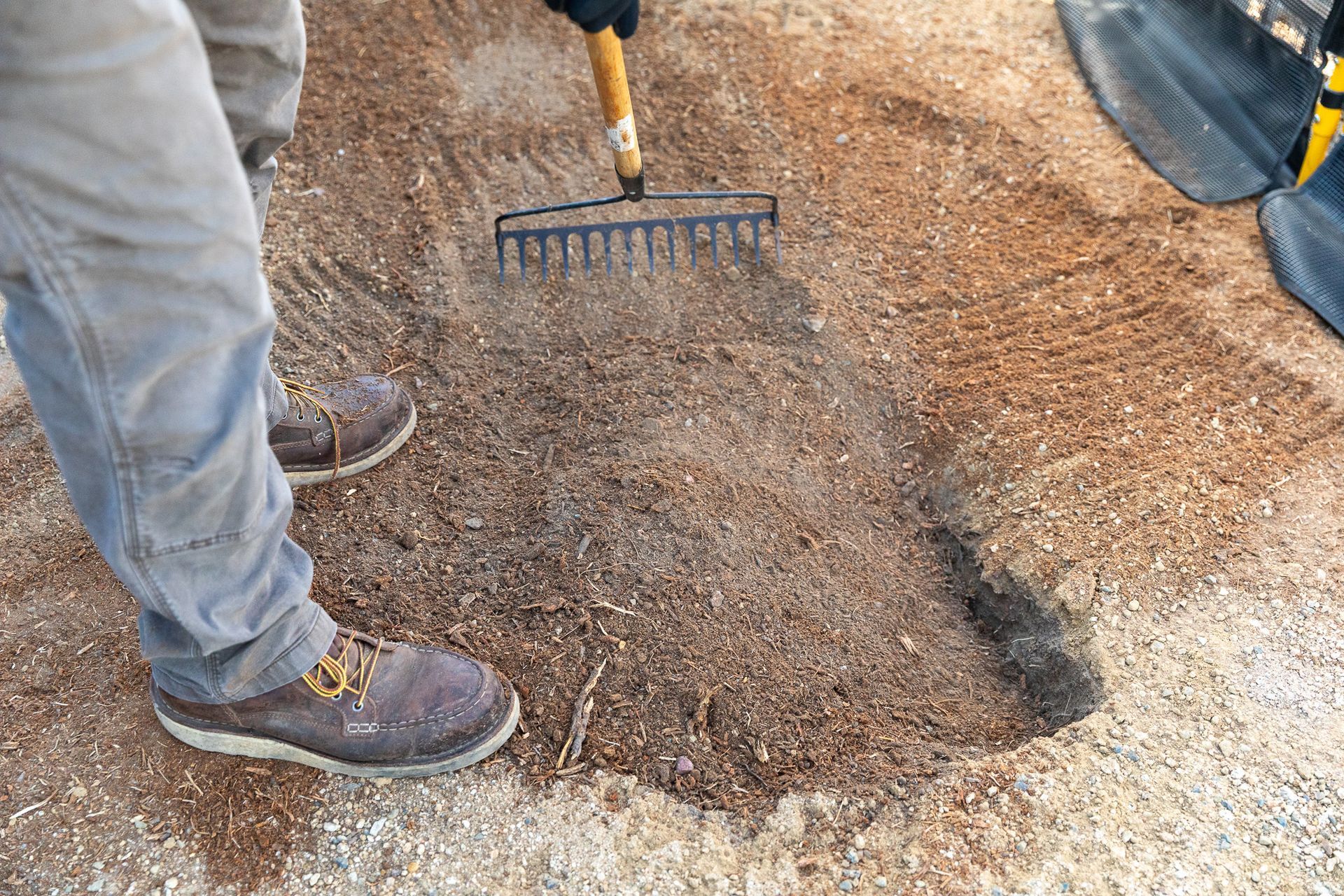 Two workers rake and spread gravel on a dirt surface outdoors.