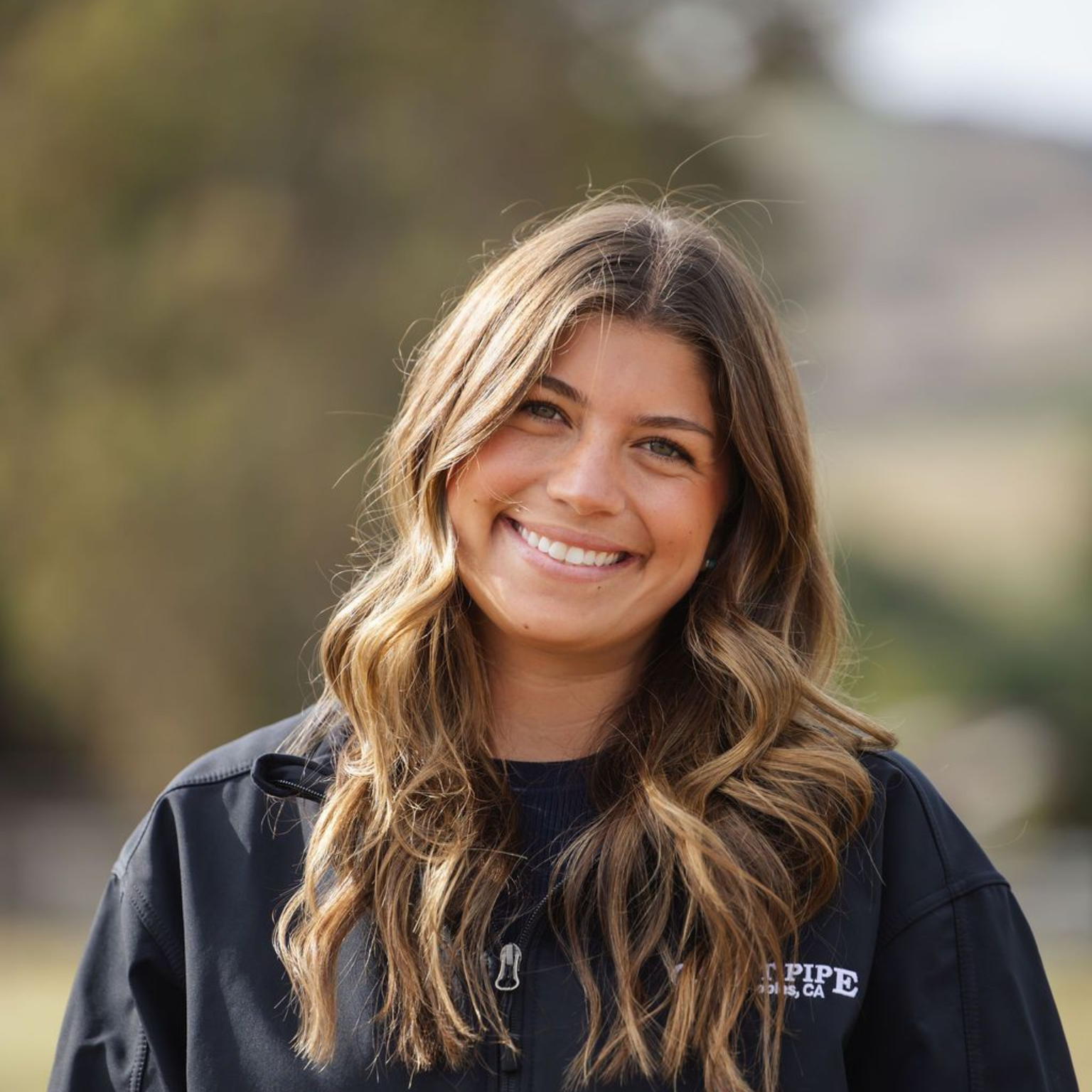 Smiling woman in a black jacket outdoors with a blurred natural background