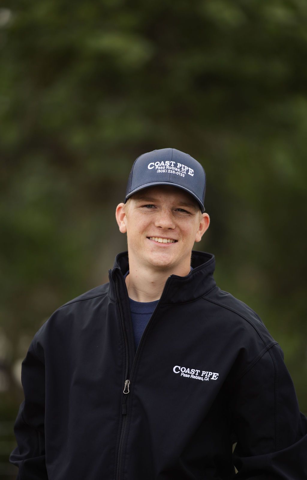 Smiling person in a black cowboy hat and jacket standing outdoors on a grassy hill