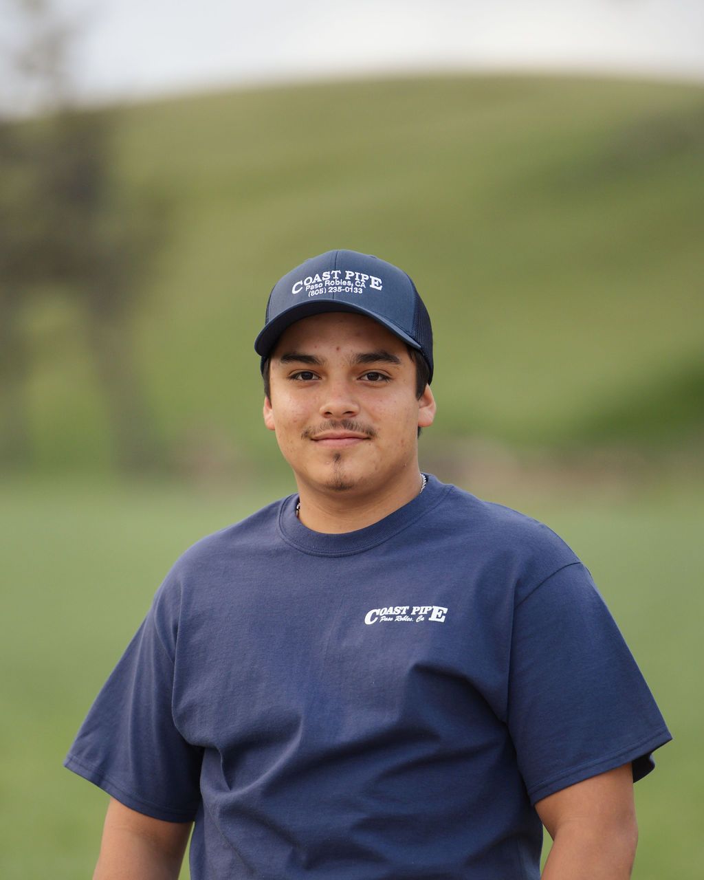 Person in a navy cap and T-shirt standing outdoors with a blurred green hillside behind them.