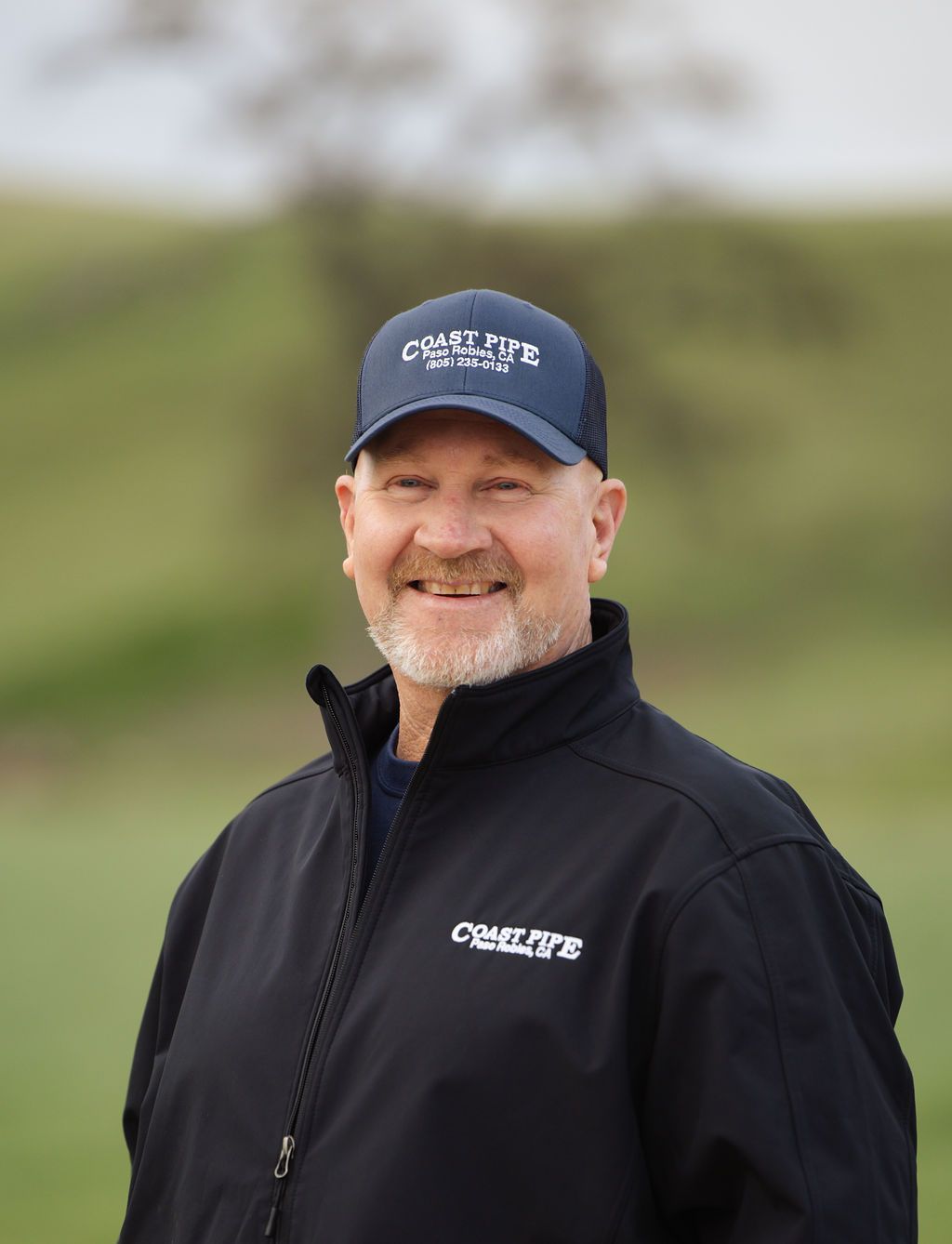 Smiling person in a navy cap and black jacket standing outdoors with a blurred green background