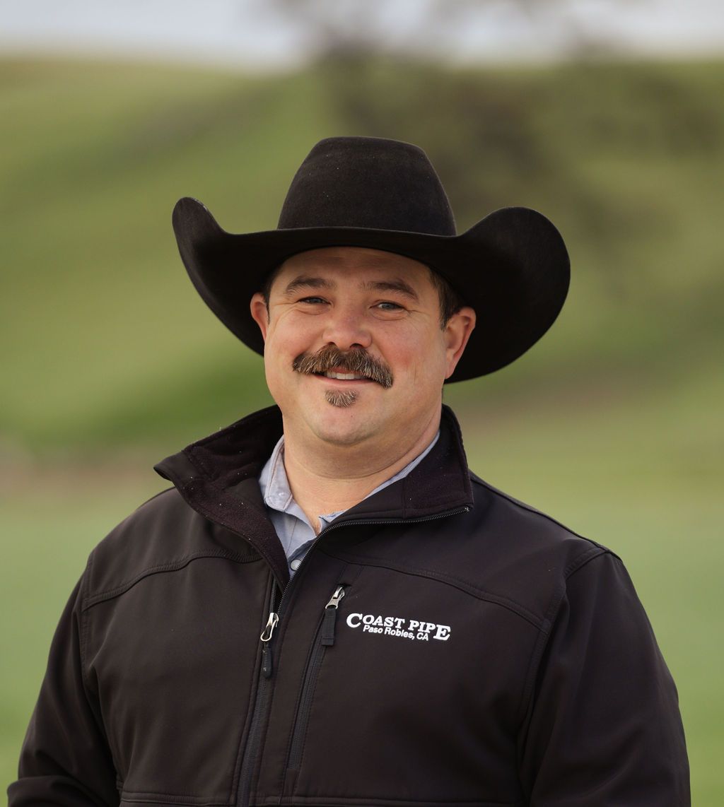 Man in a black cowboy hat and jacket standing outdoors on a grassy hillside