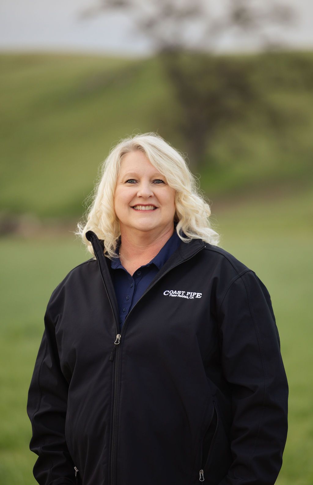A smiling woman with blonde hair, wearing a dark blue polo and black jacket, poses outdoors against a green grassy field.
