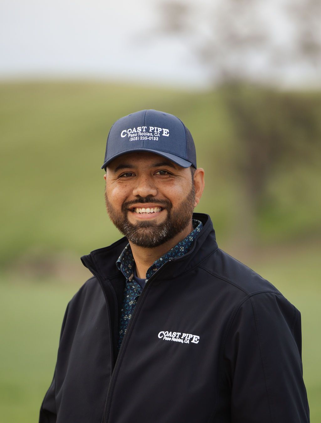 Smiling person in navy cap and jacket standing outdoors against a blurred green hillside