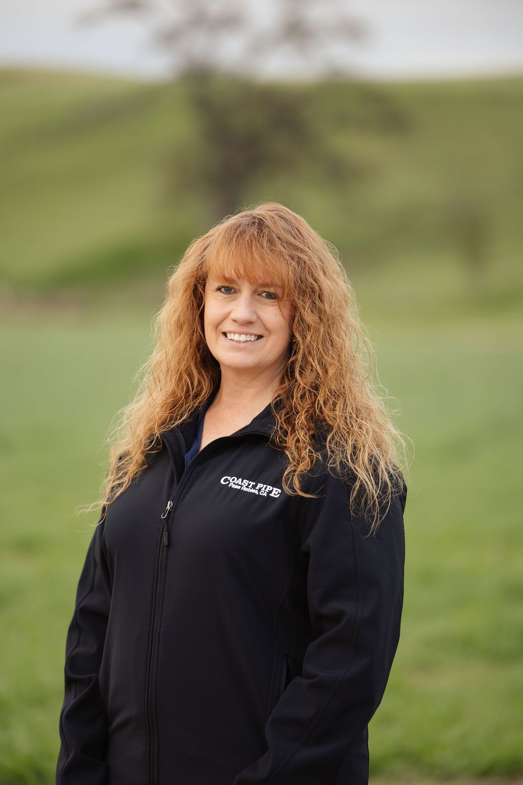 A person with long, curly auburn hair and a friendly smile, wearing a black zip-up jacket outdoors against a green field.