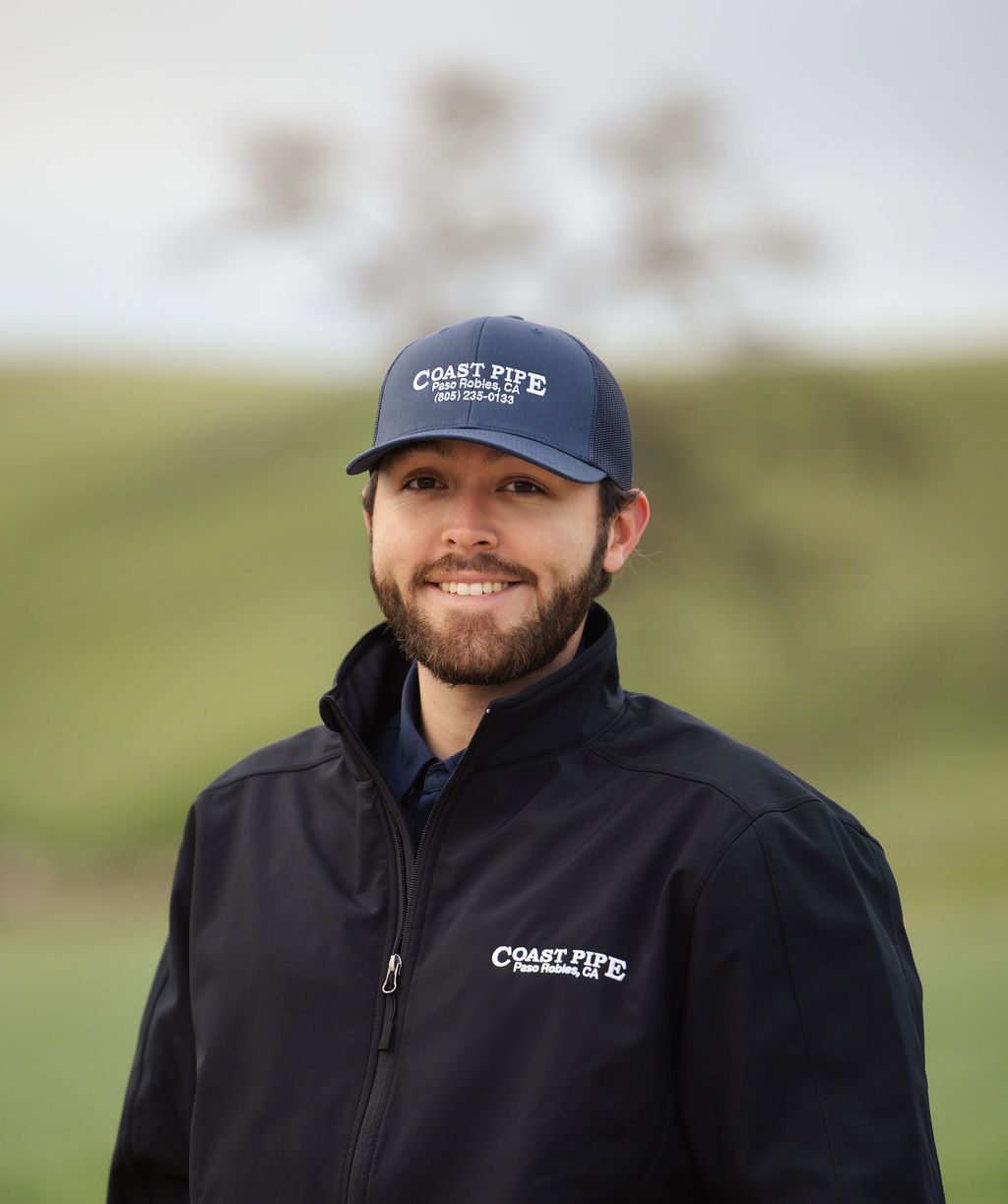 Smiling person in a navy cap and jacket standing outdoors with a blurred green field background