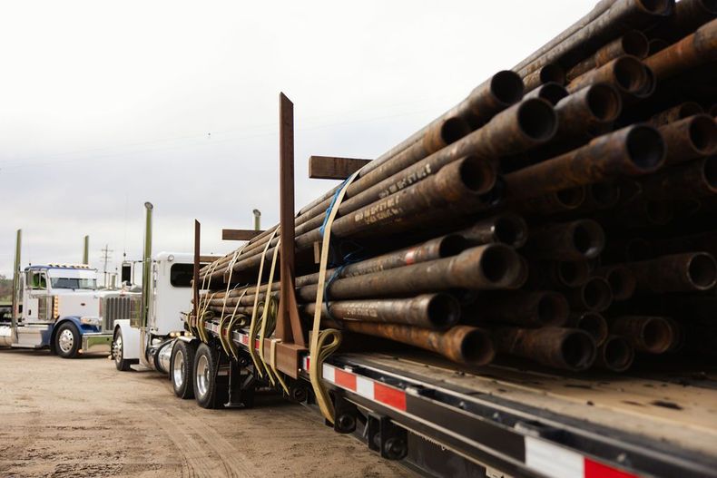 Loaded flatbed trucks hauling long stacked steel pipes in a dusty industrial yard