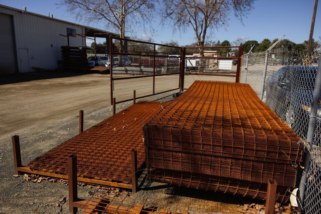 Rusty metal rebar panels stacked outdoors beside a chain-link fence and industrial building.