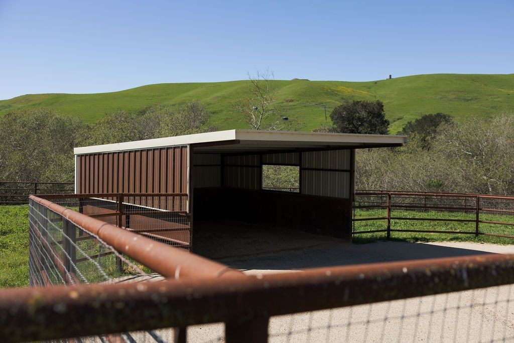 Rusty-roof cattle shelter in a fenced pasture with green hills under a blue sky
