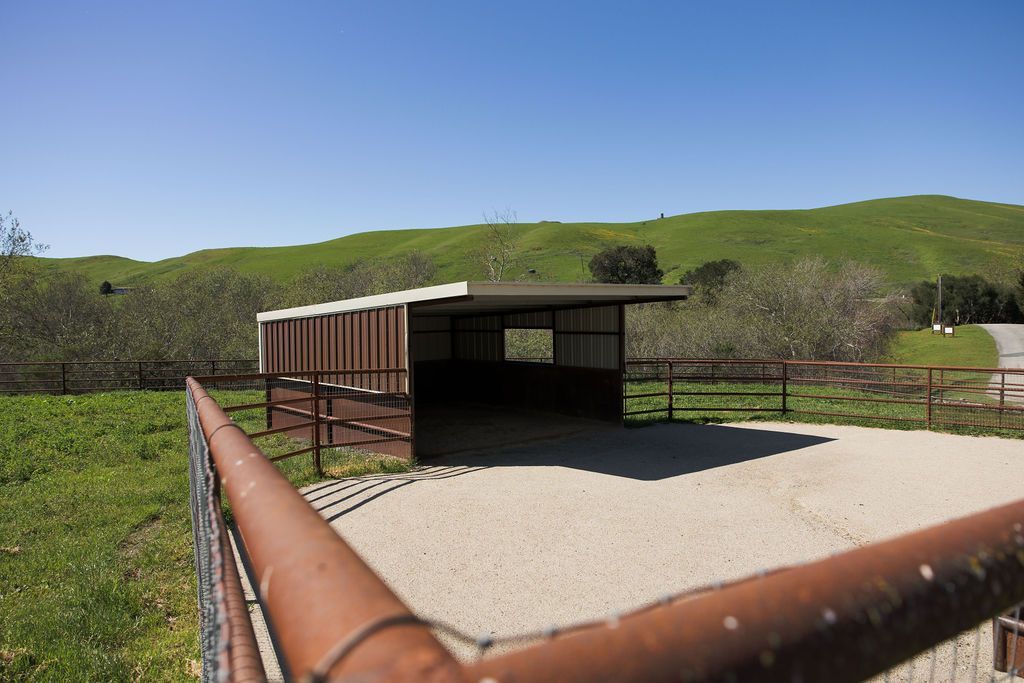 Rural shelter with rusted railings and gravel area, set against green hills under a blue sky.