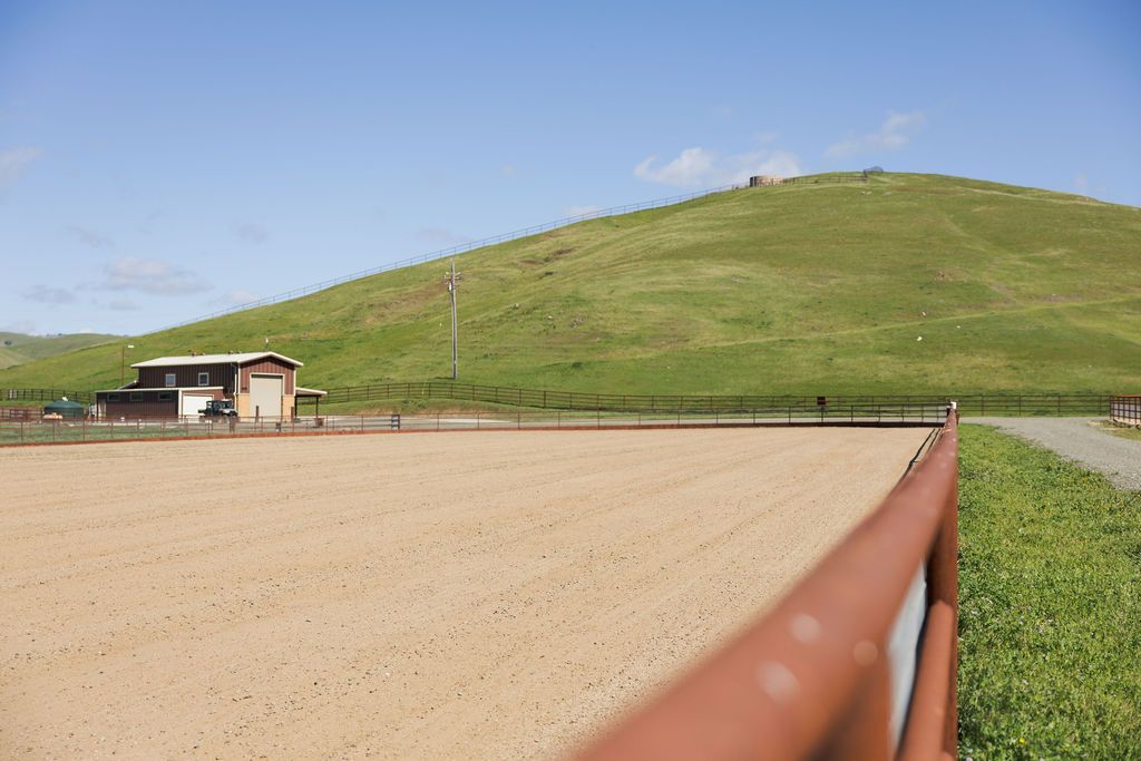 Rural dirt arena beside a grassy hill with a fence in the foreground and a small building at left.
