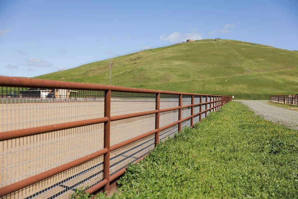 Rust-colored fence beside a gravel path with a green hillside under a blue sky