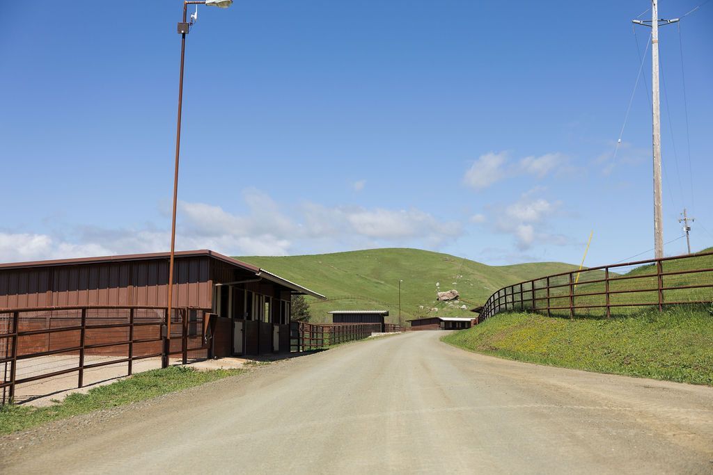 Dirt road between rural farm buildings and green hills under a clear blue sky