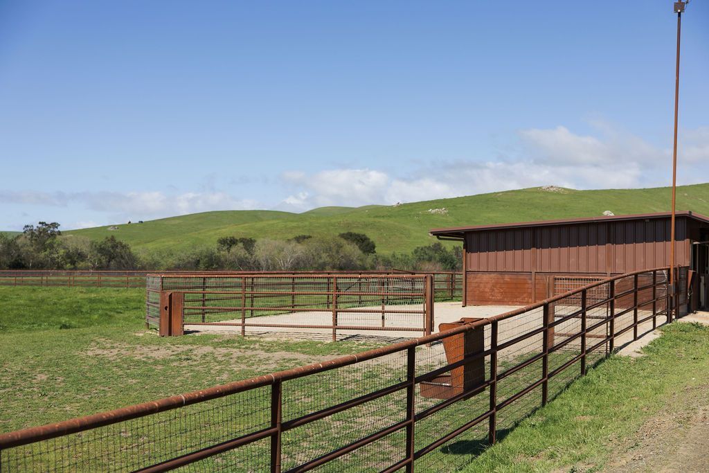 Rural horse corral with red metal fence beside a barn under a blue sky.