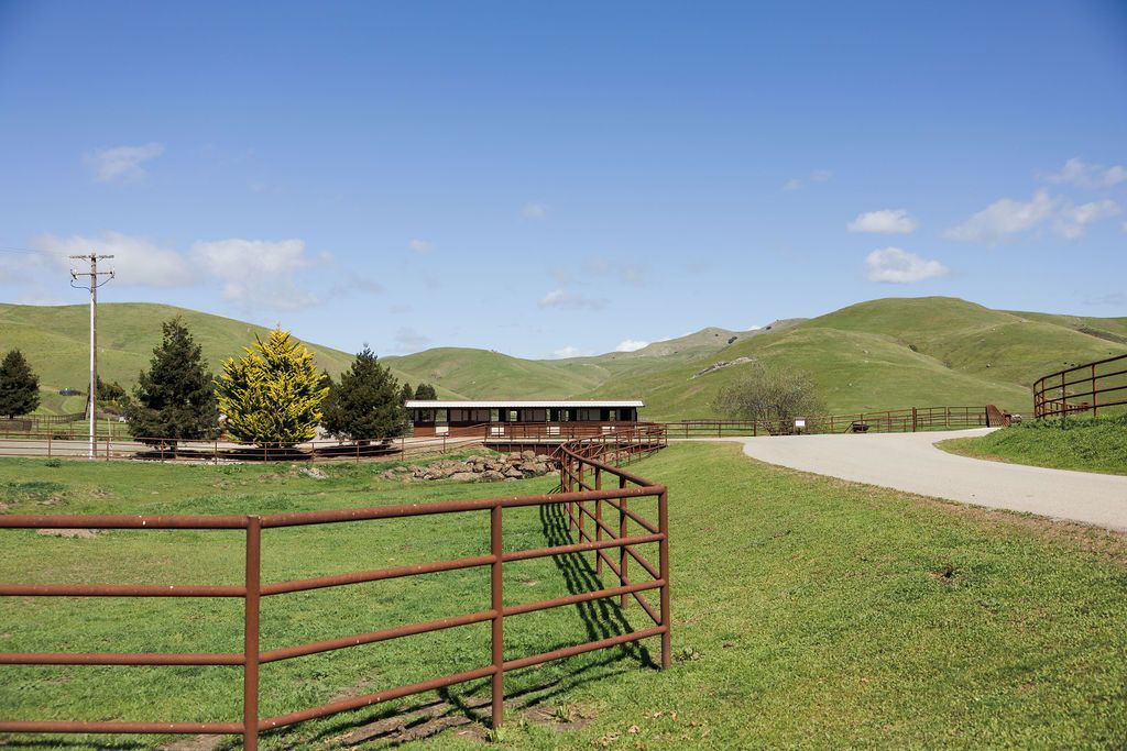 Red metal gate opens to a grassy field with rolling green hills, a small barn, and blue sky with clouds.