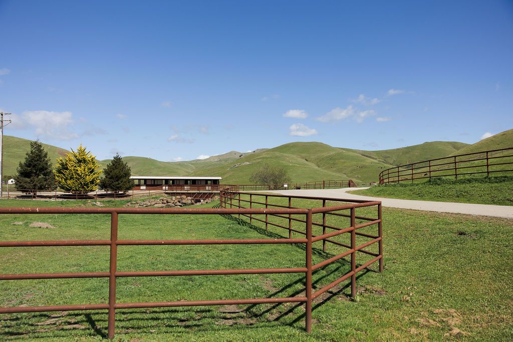 Rural pasture with red fence, gravel road, green hills, and a building under a blue sky