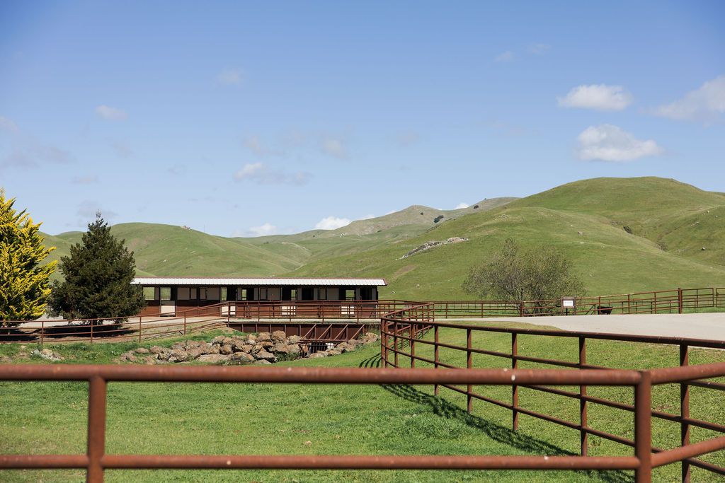Ranch fence in foreground with a barn and green hills under a blue sky