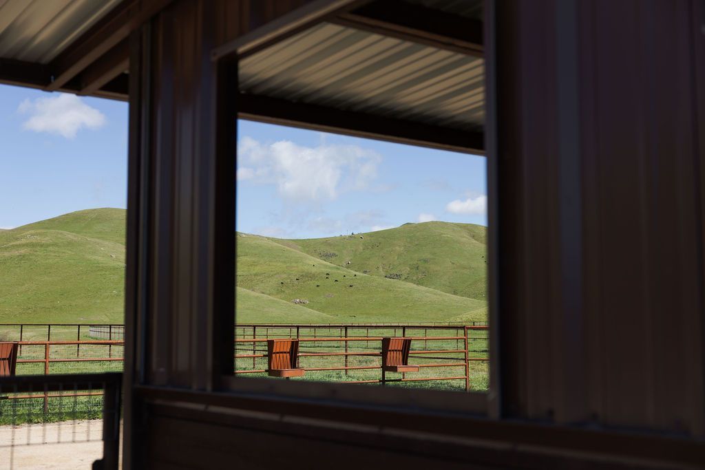 View through a window of green hills and blue sky, with a fenced porch and two chairs in the foreground