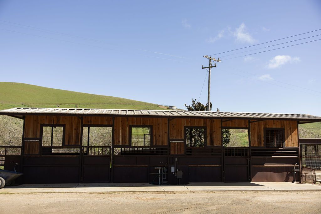 Rustic wooden building with a dark fence under a clear blue sky, beside a grassy hill