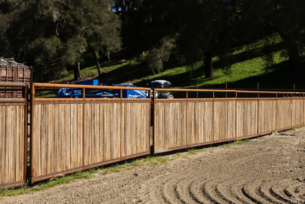 Wooden fence beside a dirt field with trees and parked blue cars beyond it