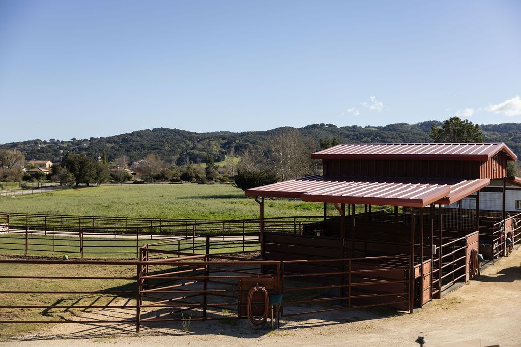 Rural horse corral with a red-roofed shelter, fenced pens, and green hills in the background