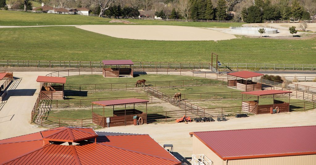 Red-roofed farm buildings and fenced animal pens in a rural landscape with green fields behind.