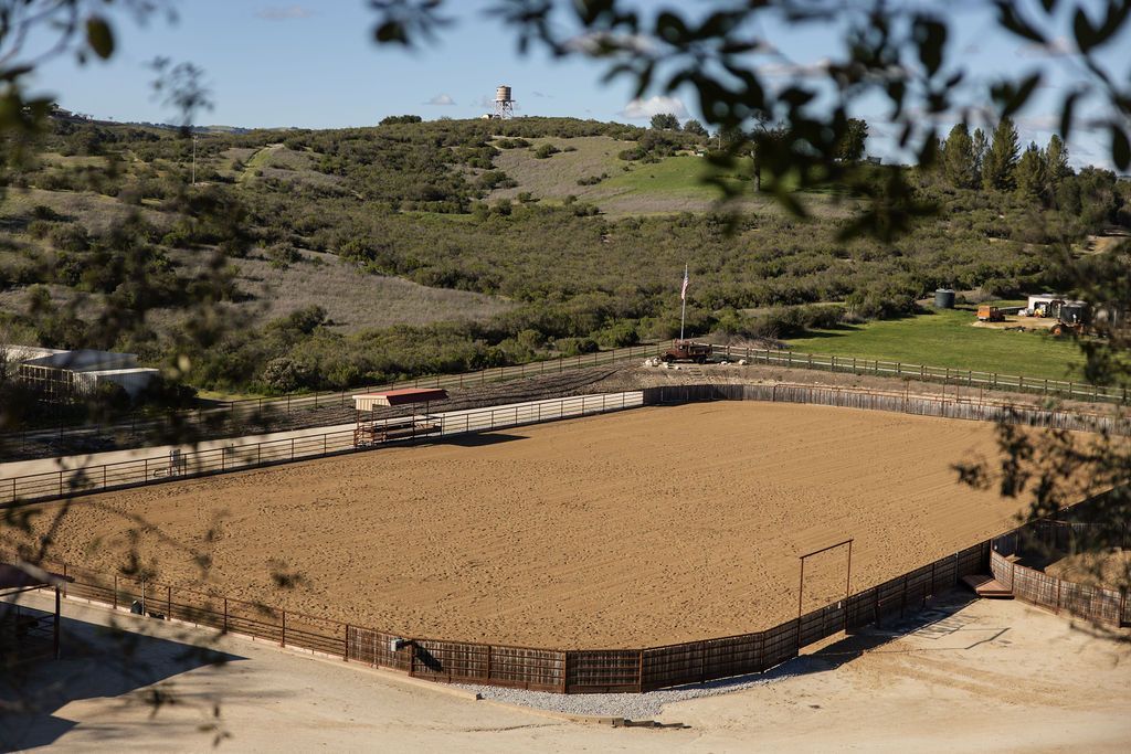 Large fenced circular dirt arena in a dry landscape, viewed from above, with trees and hills in the background