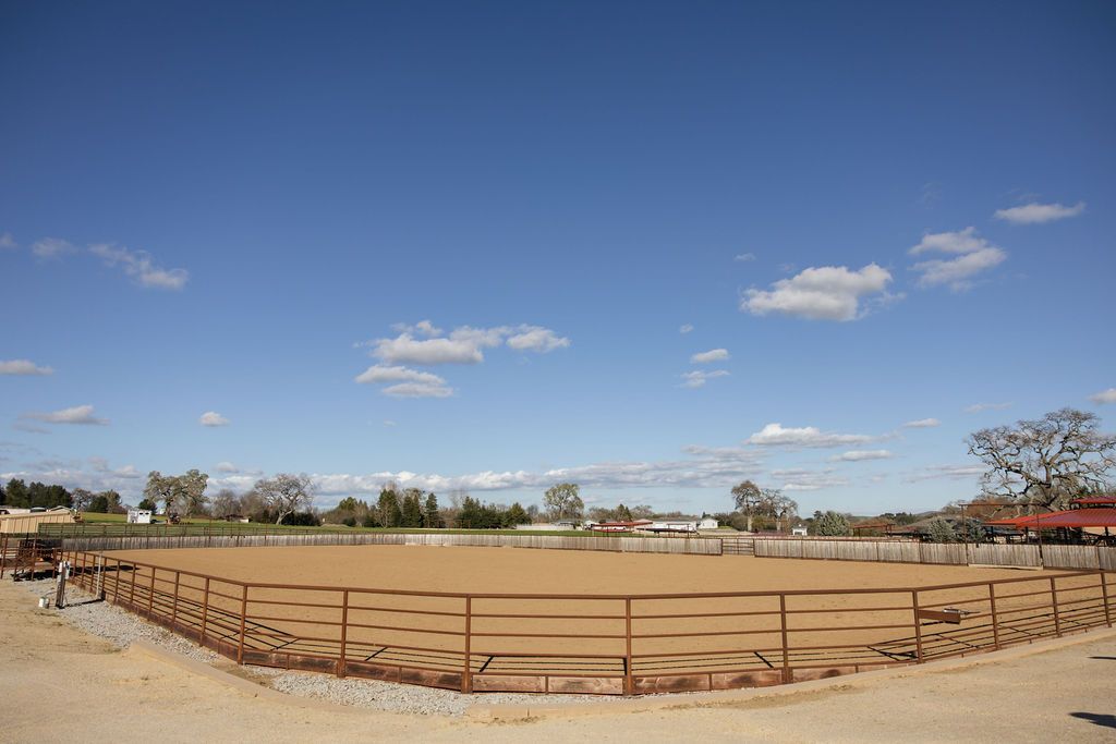 Large fenced dirt arena under a blue sky with scattered clouds, surrounded by open fields and trees.