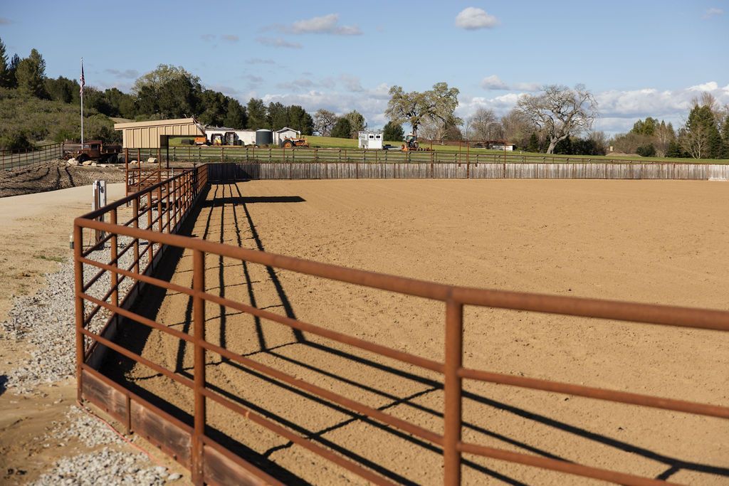 Rust-colored metal corral beside a fenced tan dirt field under a partly cloudy sky.