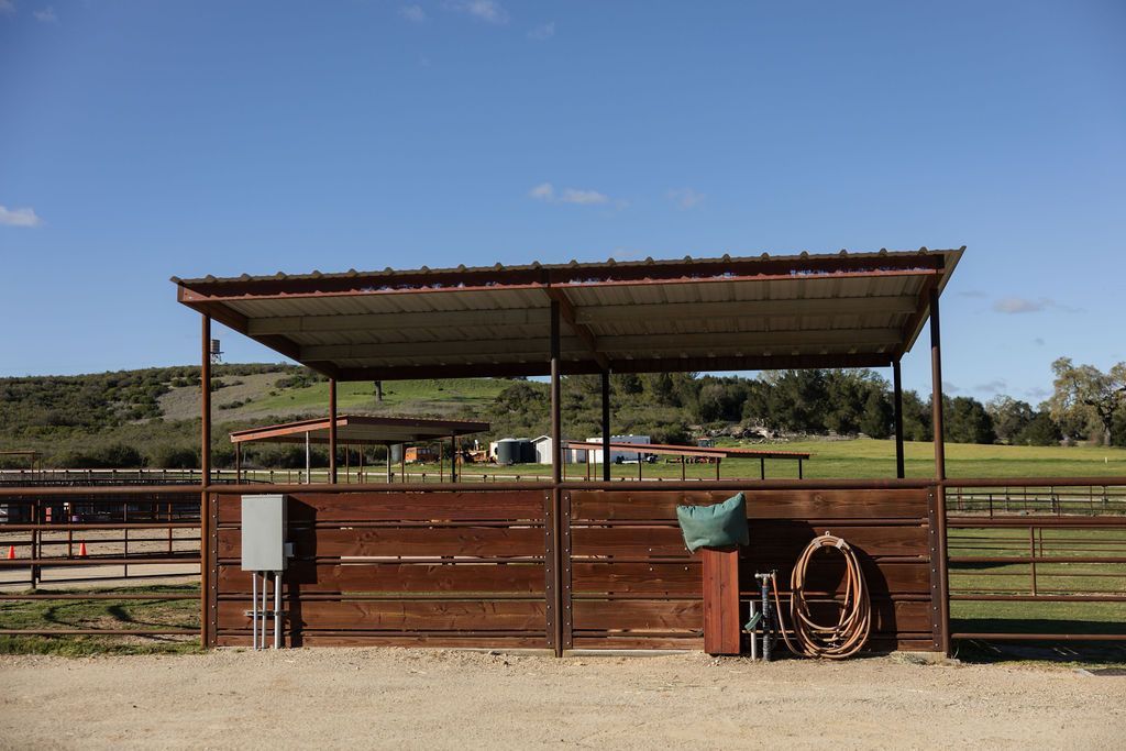Rustic horse corral with a metal-roofed shed, wooden fencing, and a coiled hose in a sunny field