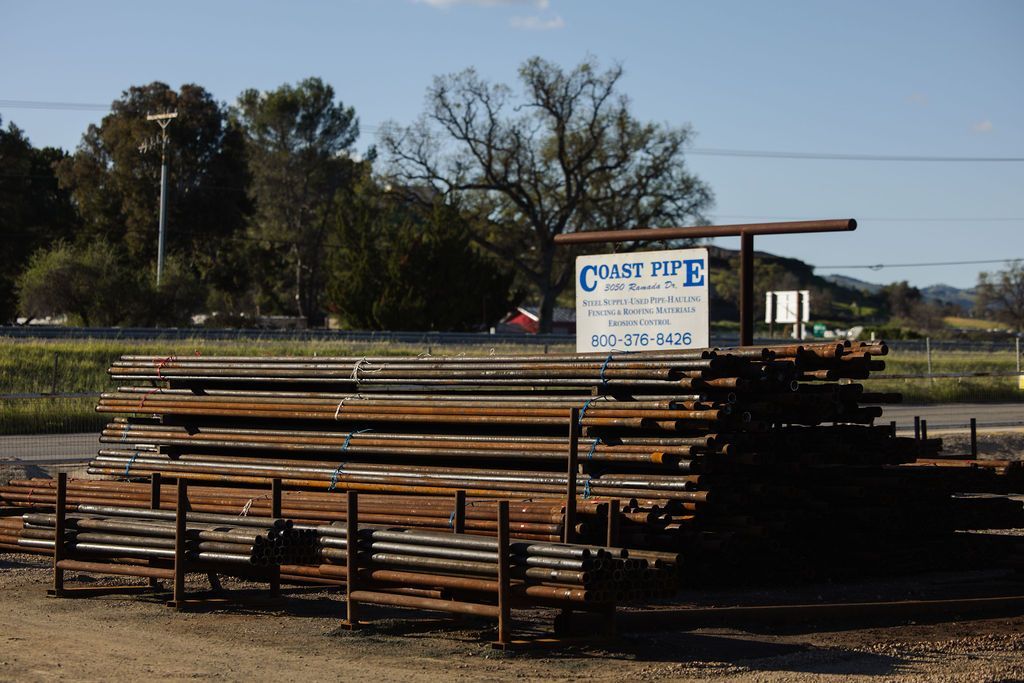 Stacked steel pipes in a yard beside a sign, with trees and open sky in the background.