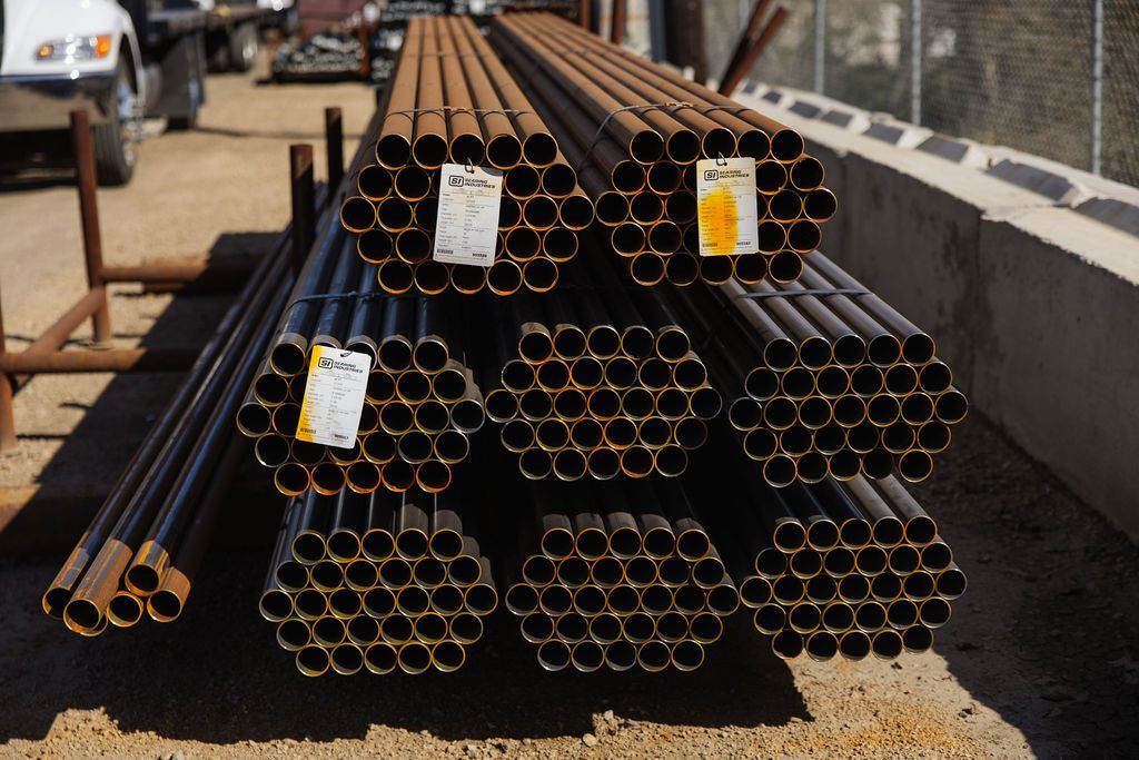 Stacks of black and rust-colored metal pipes with labels, piled outdoors on a construction site.