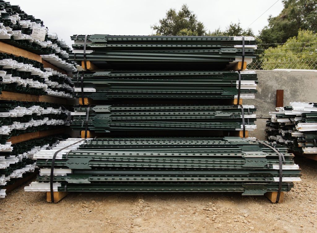 Stacks of dark green metal panels or beams stored outdoors on gravel