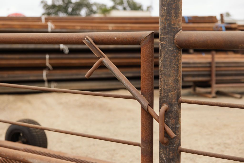Rusty metal gate latch on a farm fence with stacked pipes in the background