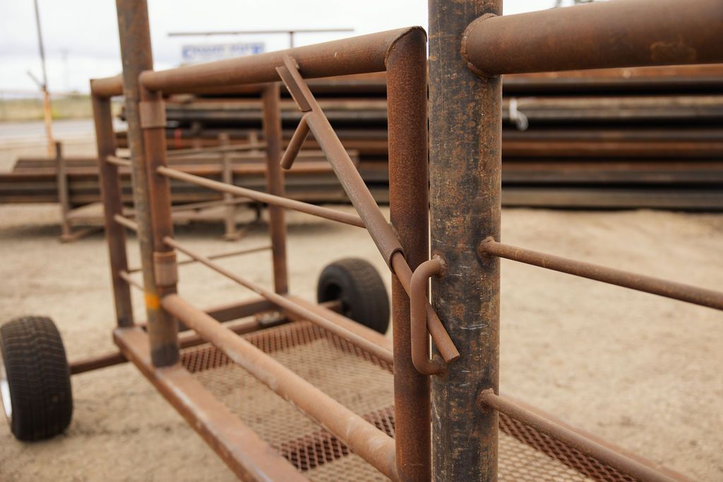 Rusty metal farm gate with wheels, leaning in a dusty yard with stacked pipes in the background