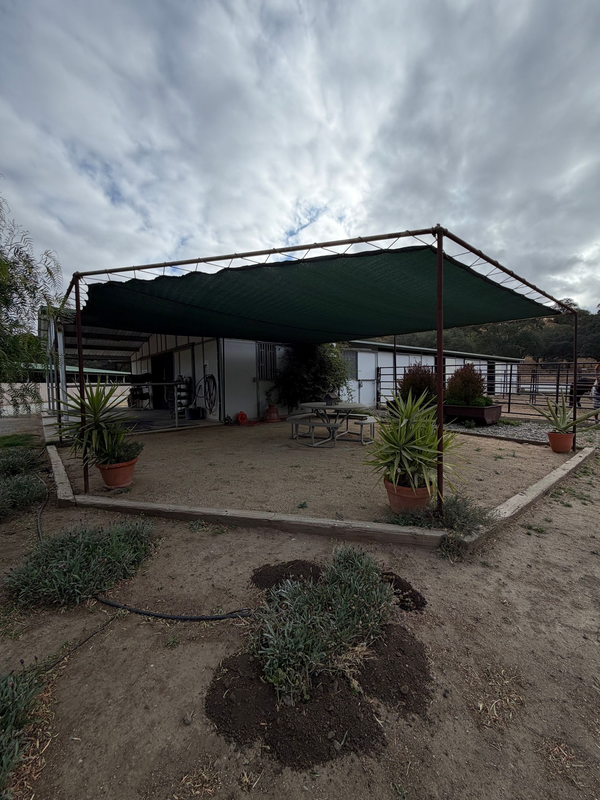 Outdoor patio with a green canopy, picnic tables, and potted plants under a cloudy sky