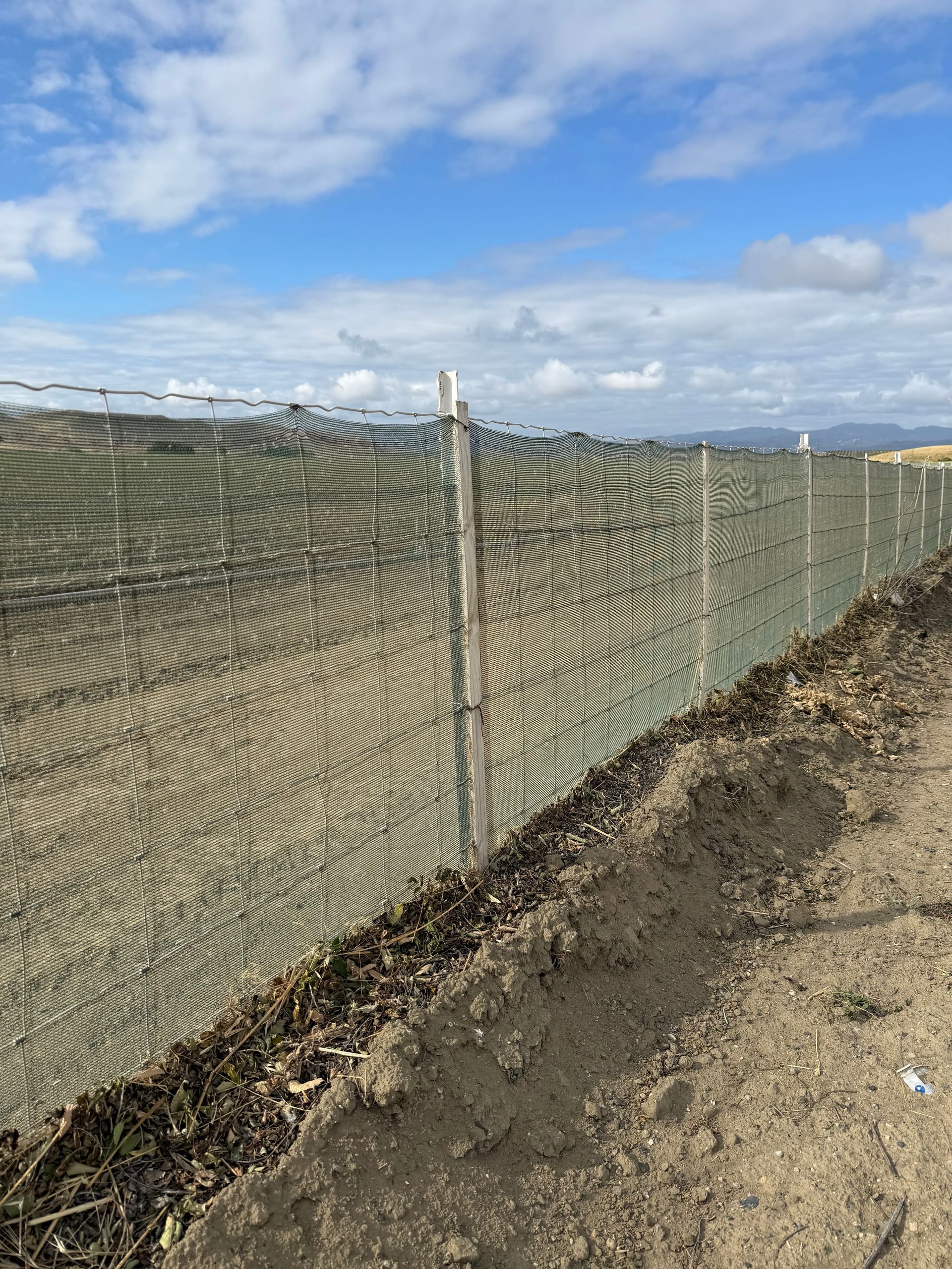 Mesh fence along a dirt path under a blue sky with clouds, overlooking open fields.