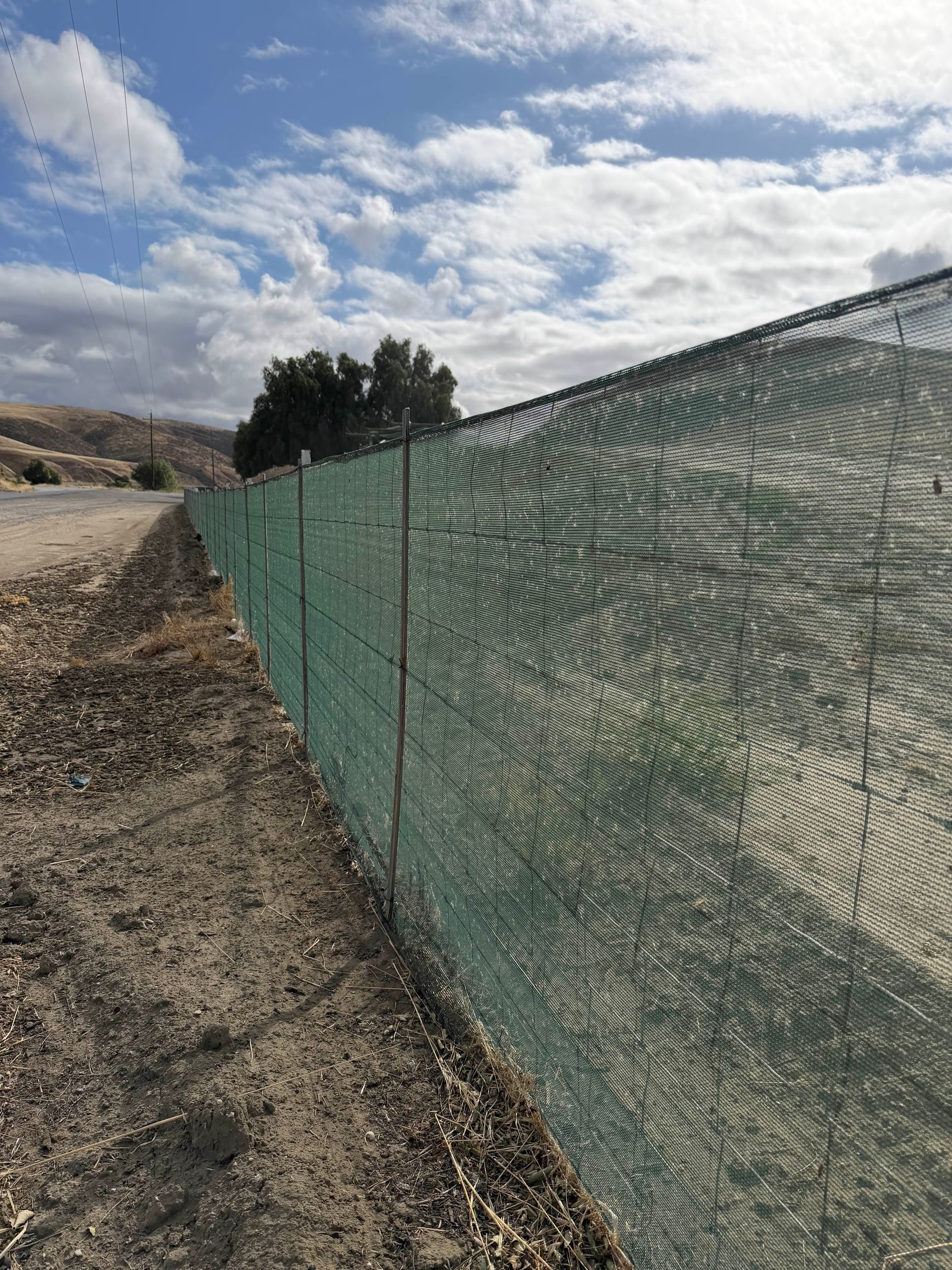 Green mesh fence along a gravel roadside under a cloudy sky, with hills in the background