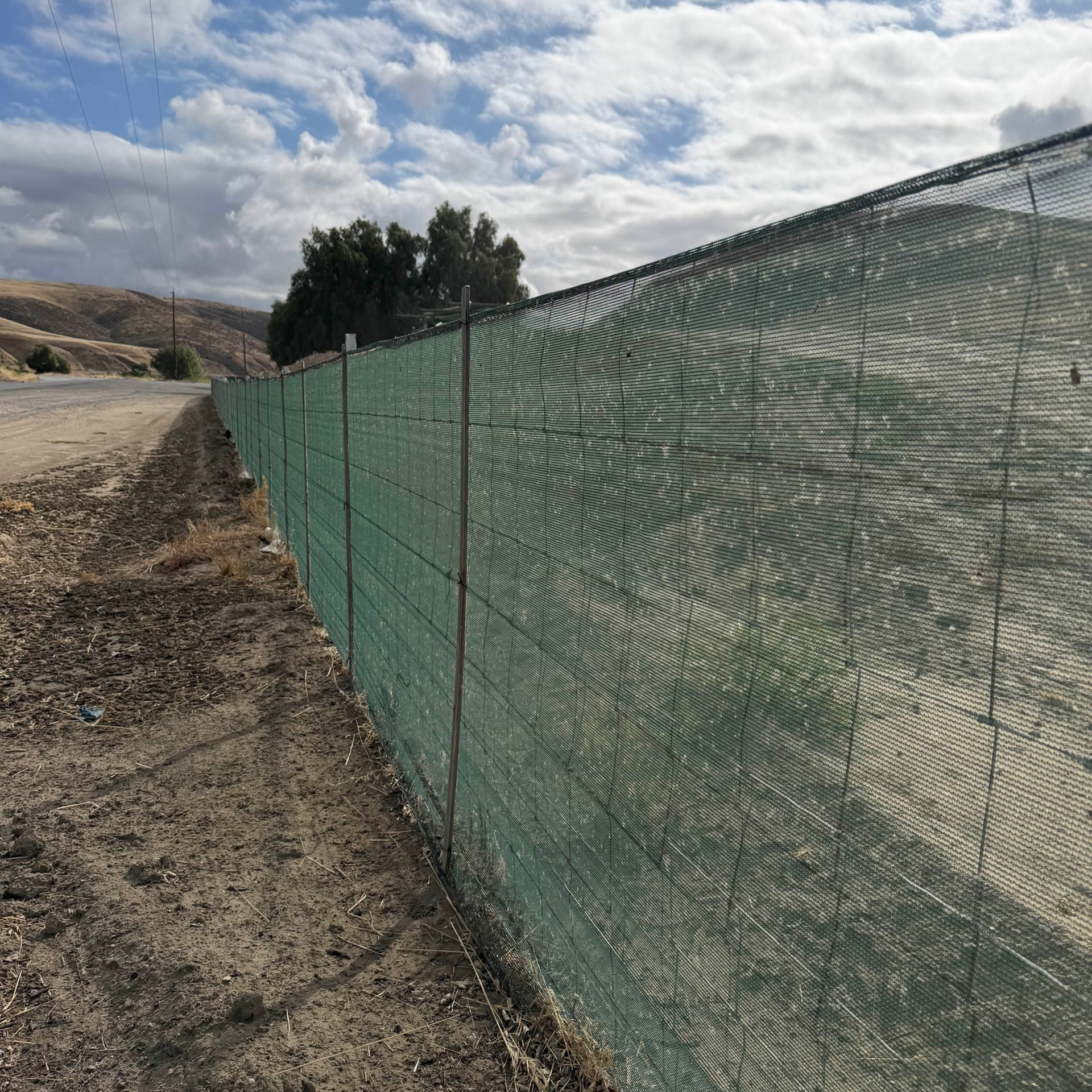 Green mesh fence along a dirt road under a cloudy sky with hills in the background