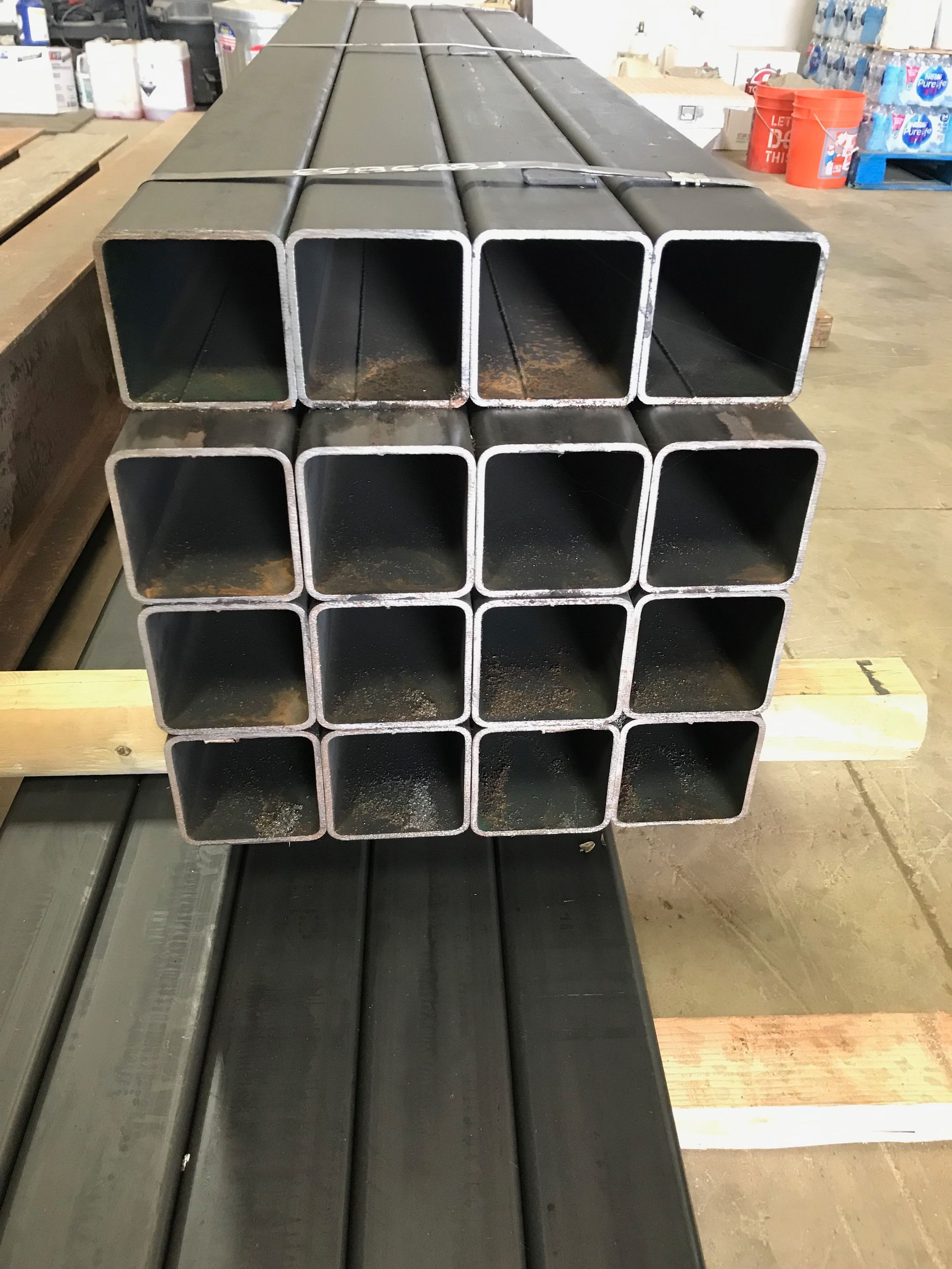 Stacked square steel tubes in a workshop, with black metal bars in the foreground.