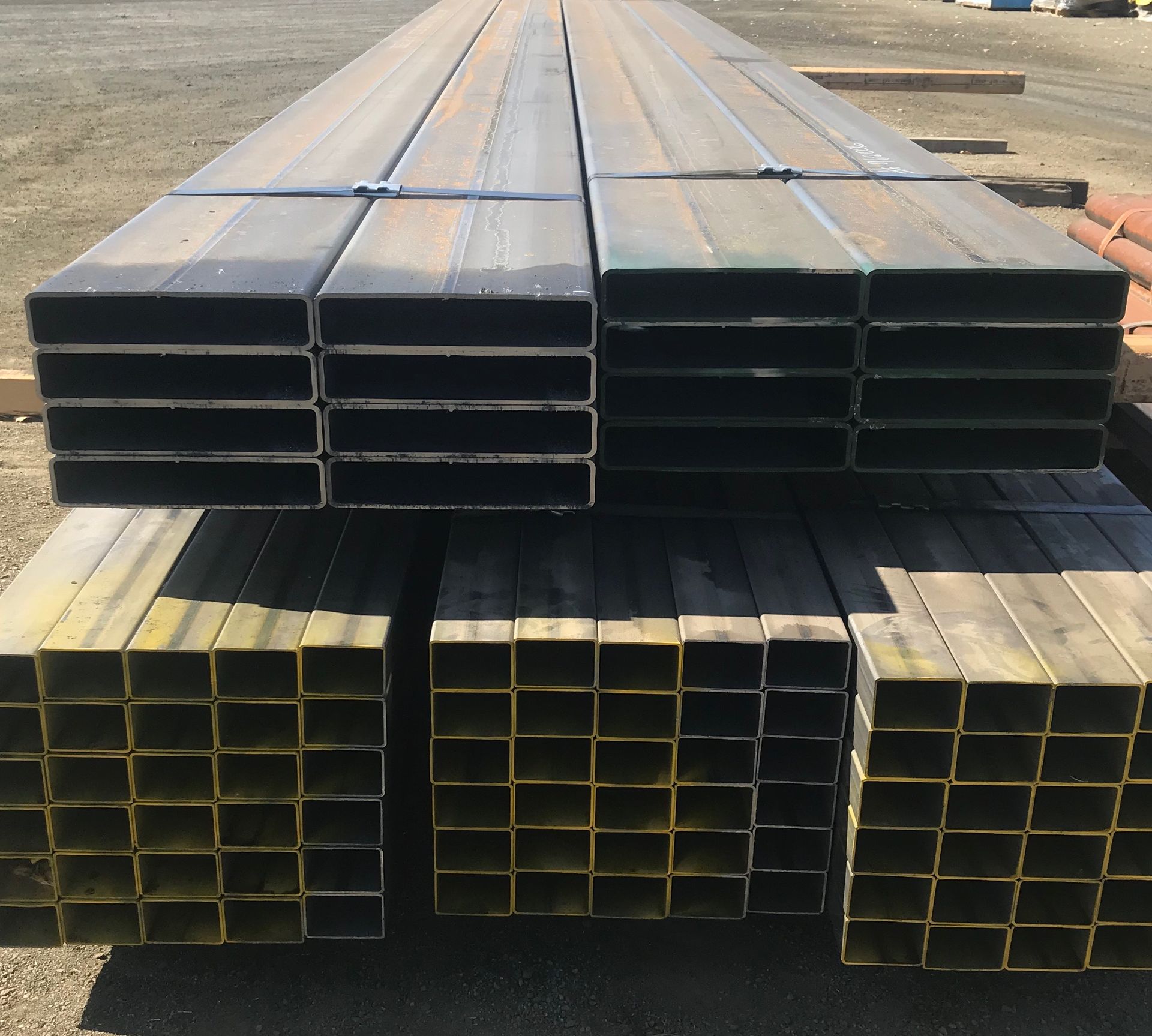 Stacks of black metal rectangular tubes and square steel pipes in an outdoor yard, viewed from the front.