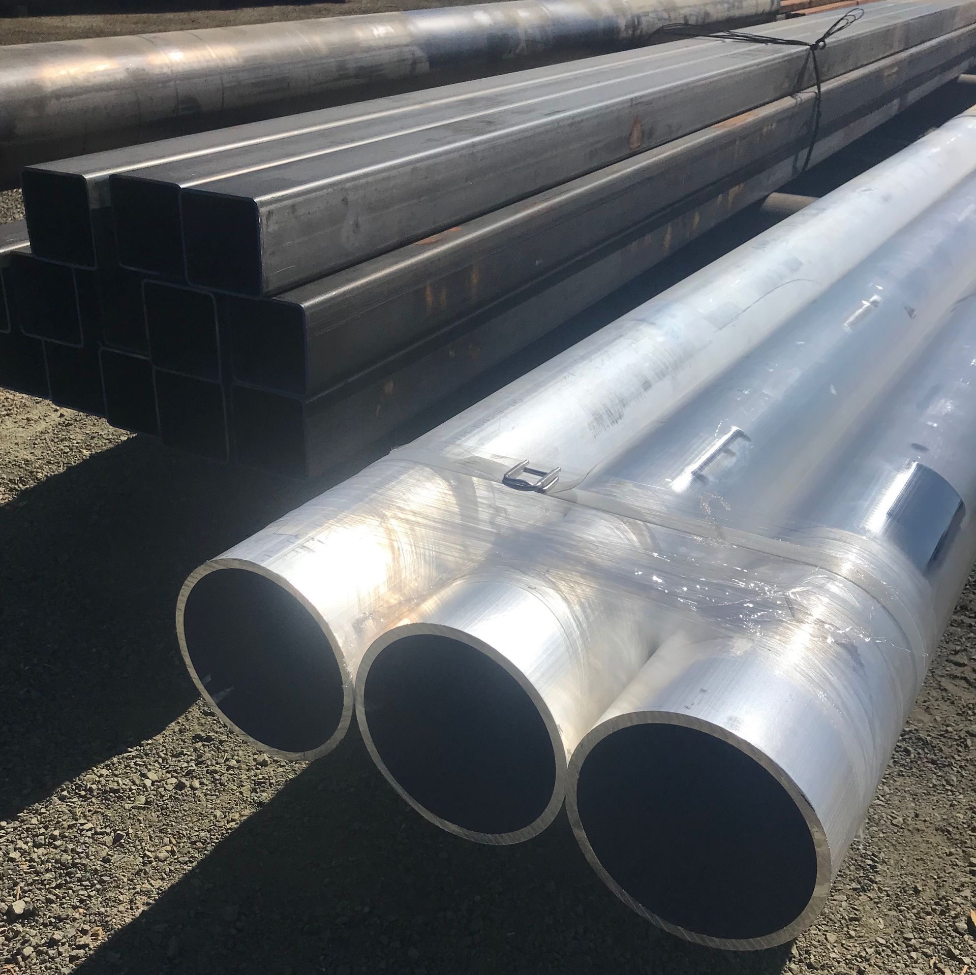 Stacks of black square steel tubes and shiny silver round pipes outdoors on gravel.