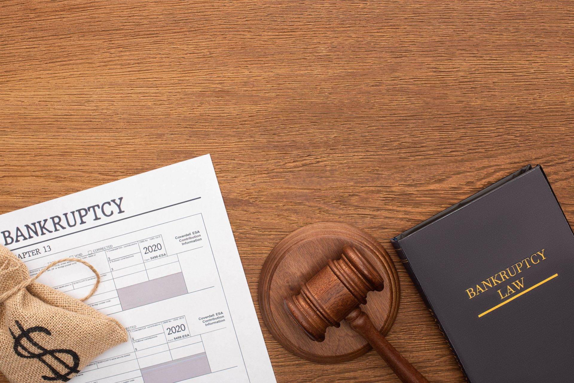 Bankruptcy paperwork, gavel, and law book on a wooden surface; a money bag is next to the paperwork.
