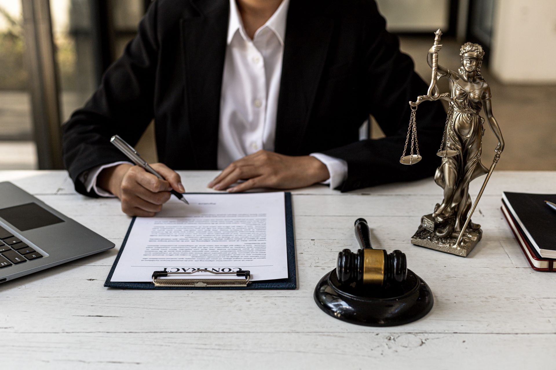 Lawyer in black suit writing at desk with clipboard, laptop, scales of justice, and gavel