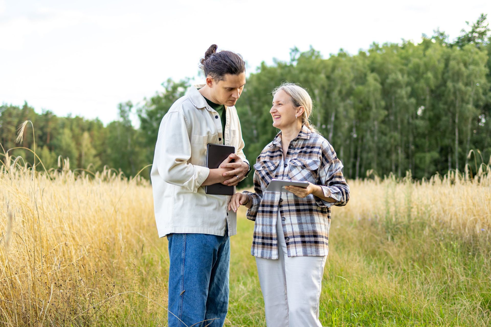 Two people smiling and talking outdoors in a grassy field, holding tablets with trees in the background