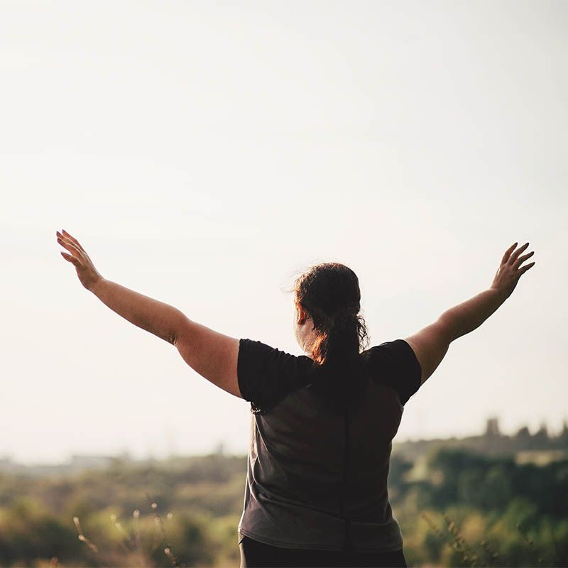 Person standing outdoors with arms raised against a bright sky