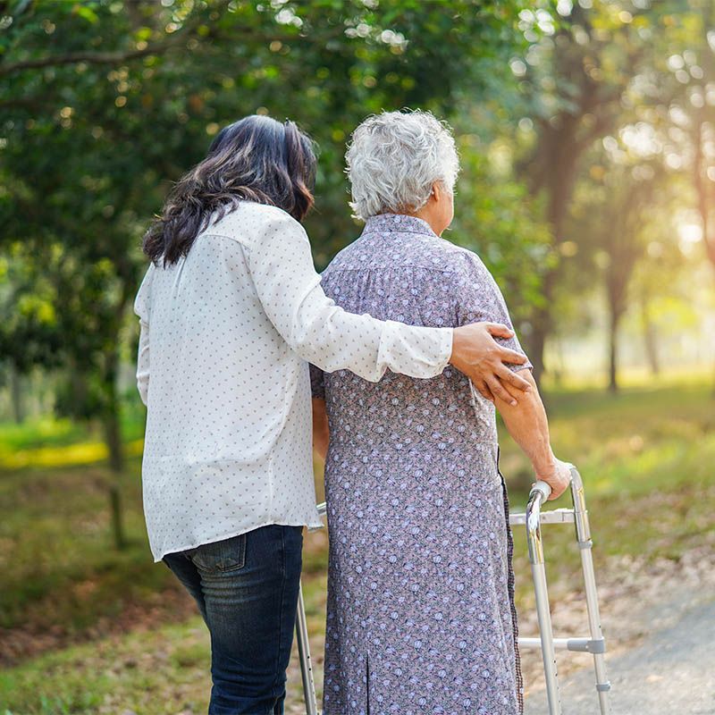 Two people walking on a sunlit park path, one supporting the other with a hand on their back and a walker