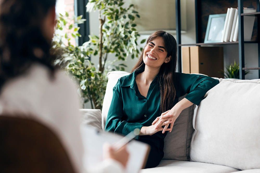 Woman smiling on a couch in a bright living room, talking with someone off camera