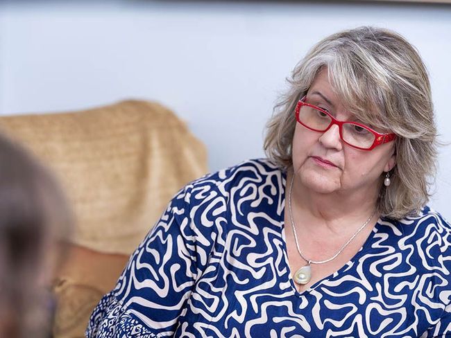 Woman in red glasses and blue patterned blouse seated on a beige sofa, looking down thoughtfully